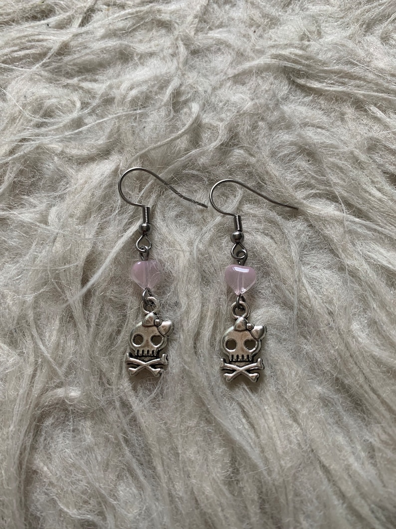 May include: A pair of silver-toned earrings. Each earring features a small, pink heart-shaped bead above a skull charm with a bow and crossbones. The earrings are suspended from a simple hook.