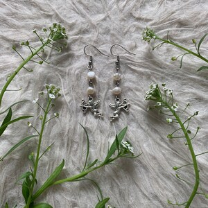 Puede incluir: Pendientes colgantes plateados con un dije de querub&iacute;n y tres perlas blancas. Los pendientes se exhiben sobre una superficie esponjosa de color gris claro, con ramitas de flores blancas y hojas verdes.