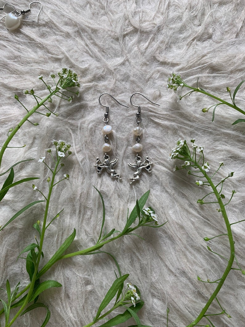 Puede incluir: Pendientes colgantes plateados con un dije de querub&iacute;n y tres perlas blancas. Los pendientes se exhiben sobre una superficie esponjosa de color gris claro, con ramitas de flores blancas y hojas verdes.