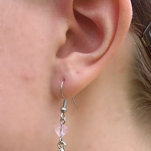 May include: Silver-tone dangle earring featuring a skull and crossbones charm with a pink heart-shaped bead. The earring is shown in a close-up view, highlighting the details of the jewelry.