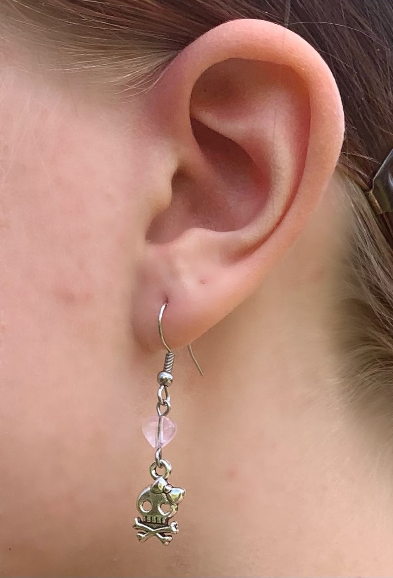 May include: Silver-tone dangle earring featuring a skull and crossbones charm with a pink heart-shaped bead. The earring is shown in a close-up view, highlighting the details of the jewelry.