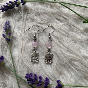 May include: A pair of silver-toned drop earrings featuring skull charms with bows and crossbones. Each earring has a pink heart-shaped bead above the skull. The earrings are displayed on a white, textured surface, with sprigs of purple lavender.