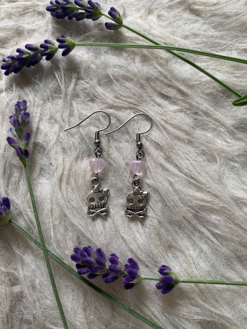 May include: A pair of silver-toned drop earrings featuring skull charms with bows and crossbones. Each earring has a pink heart-shaped bead above the skull. The earrings are displayed on a white, textured surface, with sprigs of purple lavender.