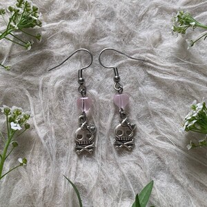 May include: A pair of dangle earrings featuring a silver skull charm with a bow and crossbones, paired with a pink heart-shaped bead. The earrings have silver hooks and are displayed on a textured, light-colored surface.