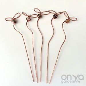 May include: Five copper wire garden stakes with a butterfly design at the top, each featuring a different colored bead. The stakes have a wavy stem and are arranged on a white surface. The brand name "onya garden" is visible in the bottom right corner.