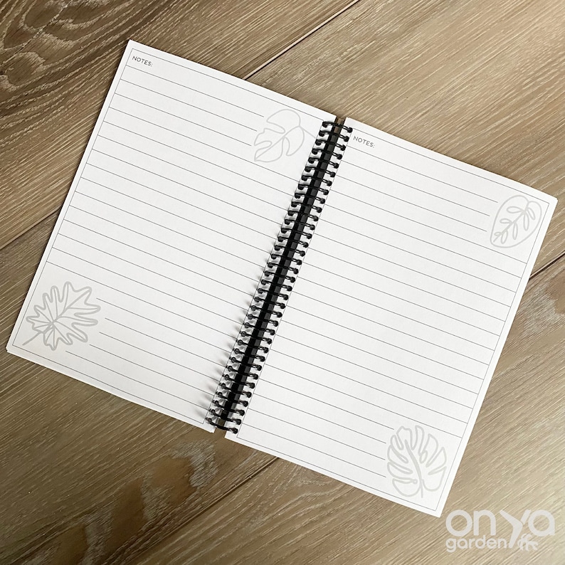 Puede incluir: Un cuaderno abierto de espiral con p&aacute;ginas blancas y l&iacute;neas negras. La portada tiene un dise&ntilde;o sencillo con una ilustraci&oacute;n de hoja gris. El cuaderno tiene la palabra "NOTES" impresa en la parte superior de cada p&aacute;gina.