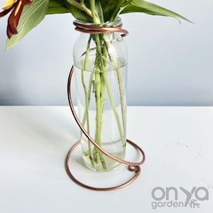 May include: A clear glass vase with a copper wire frame holding green stems and flowers. The copper wire wraps around the vase and forms a decorative base. The vase is filled with water.