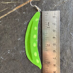 May include: Green pea pod shaped earrings with a silver hook. The earrings are approximately 7.5 cm long.