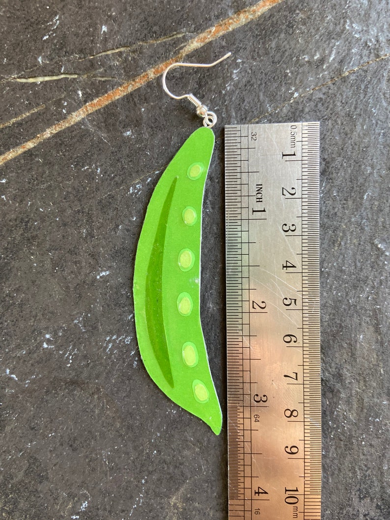 May include: Green pea pod shaped earrings with a silver hook. The earrings are approximately 7.5 cm long.