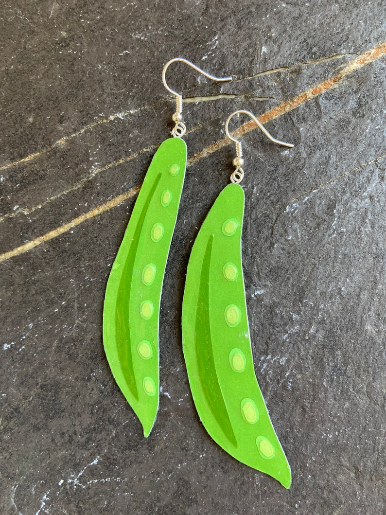 May include: A pair of green pea pod earrings with silver hooks. The earrings are made of paper and have a realistic design.