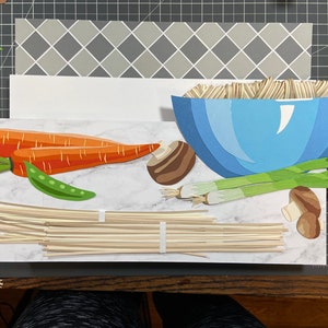 May include: A paper craft scene depicting a bowl of noodles with various ingredients, including carrots, mushrooms, green onions, and peas. The scene is set on a marble-like surface.