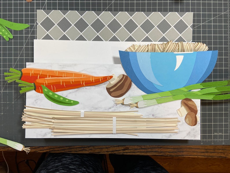 May include: A paper craft scene depicting a bowl of noodles with various ingredients, including carrots, mushrooms, green onions, and peas. The scene is set on a marble-like surface.