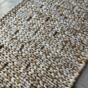 Op de afbeelding: Een geweven tapijt met een textuurpatroon in beige en bruine tinten. Het tapijt is gemaakt van natuurlijke vezels en heeft een rustieke uitstraling.