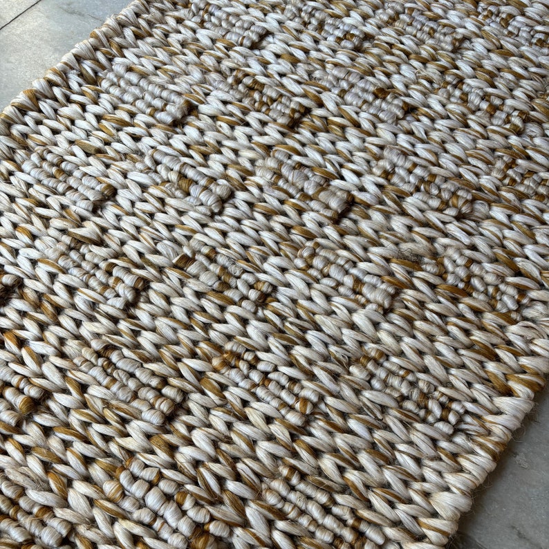 Op de afbeelding: Een geweven tapijt met een textuurpatroon in beige en bruine tinten. Het tapijt is gemaakt van natuurlijke vezels en heeft een rustieke uitstraling.