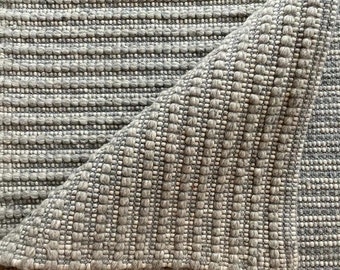May include: Close-up of a gray woven rug with a textured surface. The rug is folded in half, showing the underside of the fabric.
