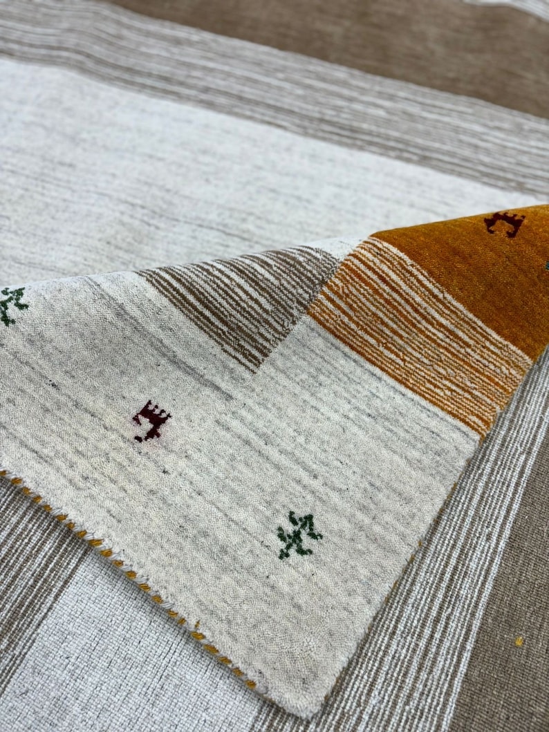May include: A white and brown striped rug with a folded corner revealing a section of the rug with orange, brown, and white stripes. The rug has small green and red embroidered designs.