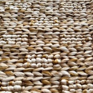 Op de afbeelding: Close-up van een geweven tapijt met een textuurpatroon. Het tapijt is gemaakt van natuurlijke vezels en heeft een beige en gouden kleurschema.