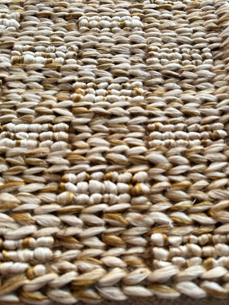 Op de afbeelding: Close-up van een geweven tapijt met een textuurpatroon. Het tapijt is gemaakt van natuurlijke vezels en heeft een beige en gouden kleurschema.