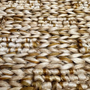 Op de afbeelding: Close-up van een geweven tapijt met een natuurlijke jute textuur. Het tapijt heeft een lichtbruine kleur met donkerbruine accenten.