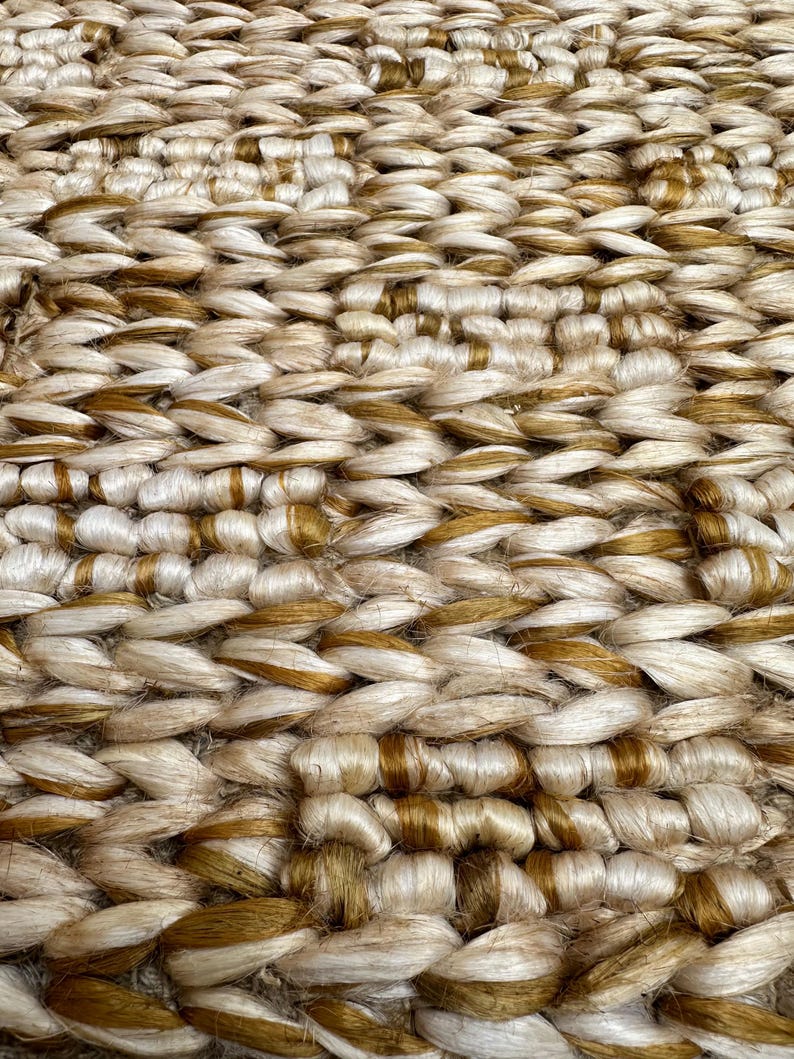 Op de afbeelding: Close-up van een geweven tapijt met een natuurlijke jute textuur. Het tapijt heeft een lichtbruine kleur met donkerbruine accenten.