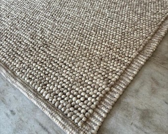 Puede incluir: Primer plano de una alfombra de lana de color marrón claro y blanco con una superficie texturizada. La alfombra tiene un borde tejido.