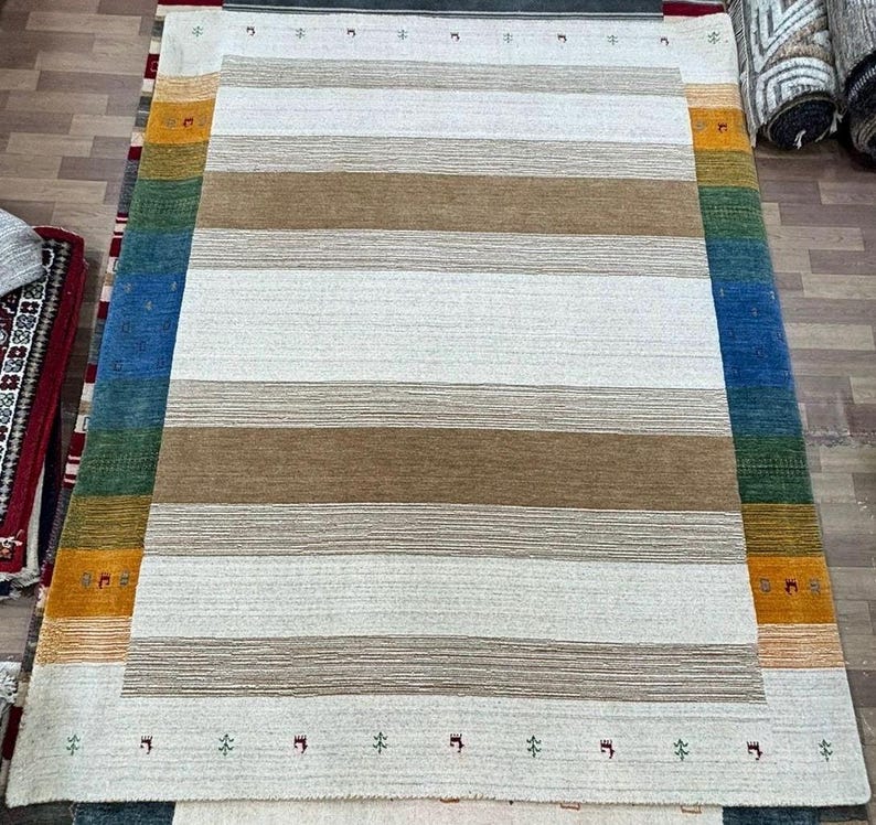 May include: A white and brown striped rug with a border of blue, green, orange, and yellow. The rug has a subtle pattern of small symbols along the border.
