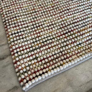 May include: A close-up of a white, cream, and multi-coloured rug made of small, round, felt balls. The rug has a subtle pattern and a light grey border.