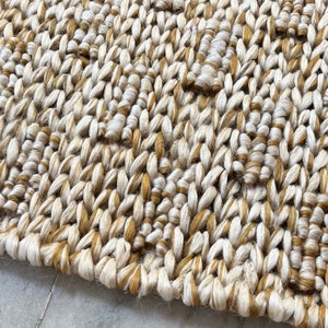 Op de afbeelding: Close-up van een geweven tapijt met een natuurlijke vezelstructuur. Het tapijt is gemaakt van een combinatie van beige en bruine garens, waardoor een subtiel patroon ontstaat.