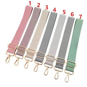 Puede incluir: Siete correas ajustables para bolsos en varios colores, incluyendo rojo, morado, beige, azul marino, gris y verde. Cada correa presenta un patrón de rayas, herrajes metálicos dorados y un cierre giratorio.