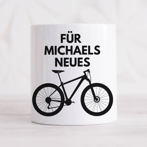 Könnte beinhalten: Weißer Keramikbecher mit schwarzer Fahrradsilhouette und dem deutschen Text "FÜR MICHAELS NEUES". Der Becher zeigt ein Mountainbike-Design, ideal für Radfahrer. Der Becher ist zylindrisch und hat einen Henkel.