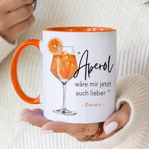 K&ouml;nnte beinhalten: Wei&szlig;e Keramiktasse mit orangefarbenem Rand. Die Tasse zeigt eine handgezeichnete Illustration eines Aperol Spritz Cocktails und den Text "Aperol" in Schwarz. Der Text unter der Illustration lautet "w&auml;re mir jetzt auch lieber" gefolgt von "Daniela".