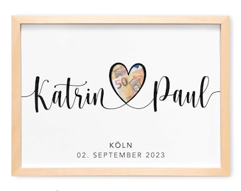 May include: A white card with black text and a heart-shaped cutout. The text reads "Katrin & Paul" with the date "02. SEPTEMBER 2023" and the location "KÖLN" below. The heart cutout is filled with a 50 Euro bill. The text "ZUM SELBER AUSSSCHNEIDEN" is below the card.