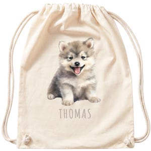 Könnte beinhalten: Ein cremefarbener Rucksack mit Kordelzug und einem aufgedruckten Bild eines flauschigen grau-weißen Welpen mit herausgestreckter Zunge. Der Name THOMAS ist in einfachen grauen Großbuchstaben direkt unter dem Welpenmotiv aufgedruckt.