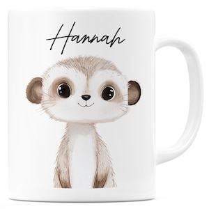 Könnte beinhalten: Weiße Keramik-Tasse mit einer Cartoon-Erdmännchen-Illustration. Das Erdmännchen ist in Braun- und Beigetönen dargestellt, mit großen Augen. Der Name "Hannah" steht in Schreibschrift über dem Erdmännchen.