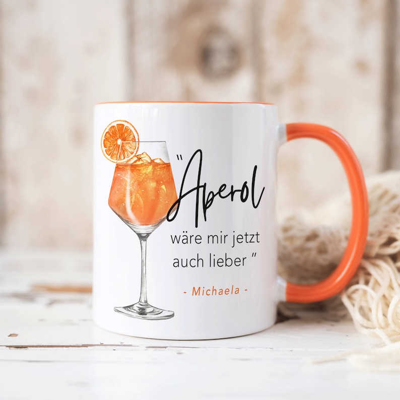 K&ouml;nnte beinhalten: Wei&szlig;e Keramiktasse mit orangefarbenem Rand und Henkel. Die Tasse zeigt eine Grafik eines Aperol Spritz Cocktails und den Text "Aperol w&auml;re mir jetzt auch lieber" - Michaela.