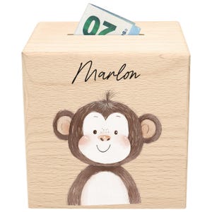 Könnte beinhalten: Eine Holzspardose mit einem Cartoon-Affen und dem Namen "Marlon" in Schreibschrift. Ein 20-Euro-Schein steckt im Schlitz. Die Box ist aus hellem Holz gefertigt.