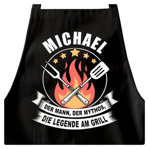 Könnte beinhalten: Schwarze Schürze mit weißem Aufdruck. Der Aufdruck zeigt den Namen "MICHAEL", eine gekreuzte Gabel und einen Pfannenwender, Flammen und Sterne. Darunter steht der Text "DER MANN, DER MYTHOS, DIE LEGENDE AM GRILL."