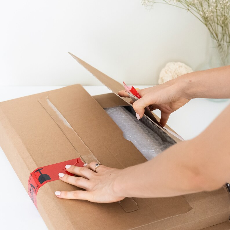 May include: A person opening a brown cardboard box with red tape. The box is filled with bubble wrap and a product is visible inside.