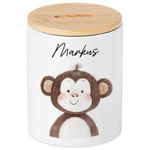 Könnte beinhalten: Weiße Keramik-Spardose mit Bambusdeckel. Die Spardose zeigt eine Cartoon-Affen-Illustration und den Namen "Markus" in schwarzer Schrift. Der Deckel hat einen Schlitz zum Einwerfen von Geld.