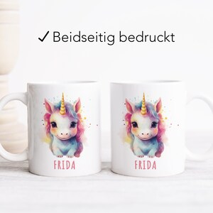 May include: Two white ceramic mugs featuring a colourful unicorn illustration and the name "FRIDA" printed on each mug. The mugs are on a white wooden surface.