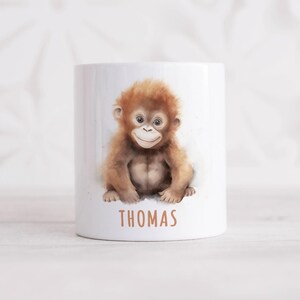 Könnte beinhalten: Weiße Keramiktasse mit einer Aquarell-Illustration eines Baby-Orang-Utans. Die Tasse trägt den Namen "THOMAS" auf dem Boden.