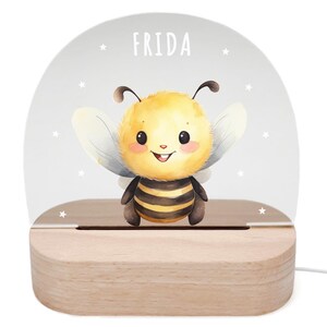 Könnte beinhalten: Ein weißes Acryl-Nachtlicht mit einer niedlichen Cartoon-Bienen-Illustration und dem Namen "FRIDA" darauf gedruckt. Das Licht sitzt auf einem Holzsockel.