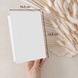 May include: A blank white card measuring 10.5 inches by 14.8 inches. The card is held in a hand against a background of beige fabric and a dried pampas grass arrangement.