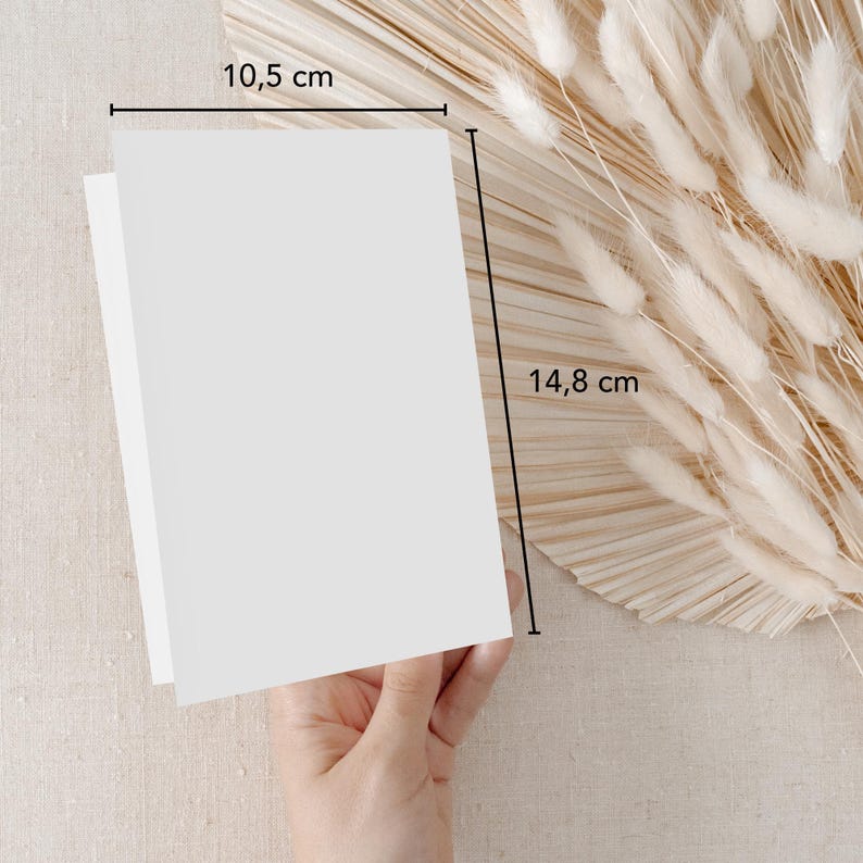 May include: A blank white card measuring 10.5 inches by 14.8 inches. The card is held in a hand against a background of beige fabric and a dried pampas grass arrangement.