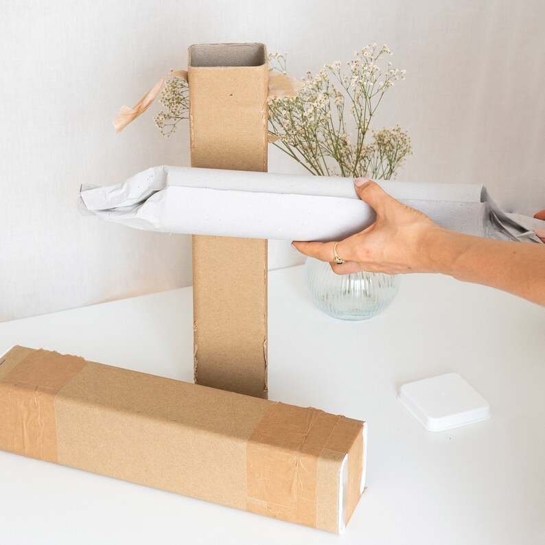 May include: Two brown cardboard boxes, one tall and one long, are stacked on a white surface. A person is holding a white paper tube over the top of the boxes. The paper tube is partially unrolled. A small white square box is in the background.