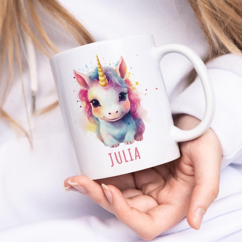 May include: White ceramic mug featuring a colourful illustration of a unicorn with a pink, yellow and blue mane and a gold horn. The mug has the name "Julia" printed in pink lettering on the bottom.