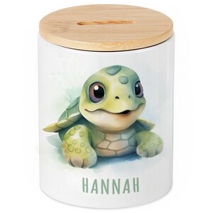 Könnte beinhalten: Weiße Keramik-Spardose mit Bambusdeckel. Sie zeigt eine Aquarell-Illustration einer lächelnden grünen Schildkröte. Der Name "HANNAH" ist in grünen Buchstaben auf der Vorderseite aufgedruckt. Die Spardose ist zum Sparen von Geld bestimmt.
