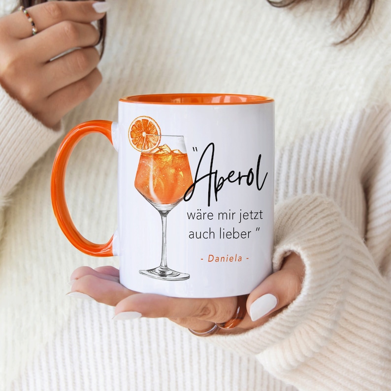 K&ouml;nnte beinhalten: Wei&szlig;e Keramiktasse mit orangefarbenem Rand. Die Tasse zeigt eine handgezeichnete Illustration eines Aperol Spritz Cocktails und den Text "Aperol" in Schwarz. Der Text unter der Illustration lautet "w&auml;re mir jetzt auch lieber" gefolgt von "Daniela".