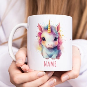 May include: White ceramic mug featuring a colourful illustration of a unicorn with a gold horn. The unicorn is sitting on a watercolour background with pink, blue and yellow splatters. The text "NAME" is printed in pink below the unicorn.