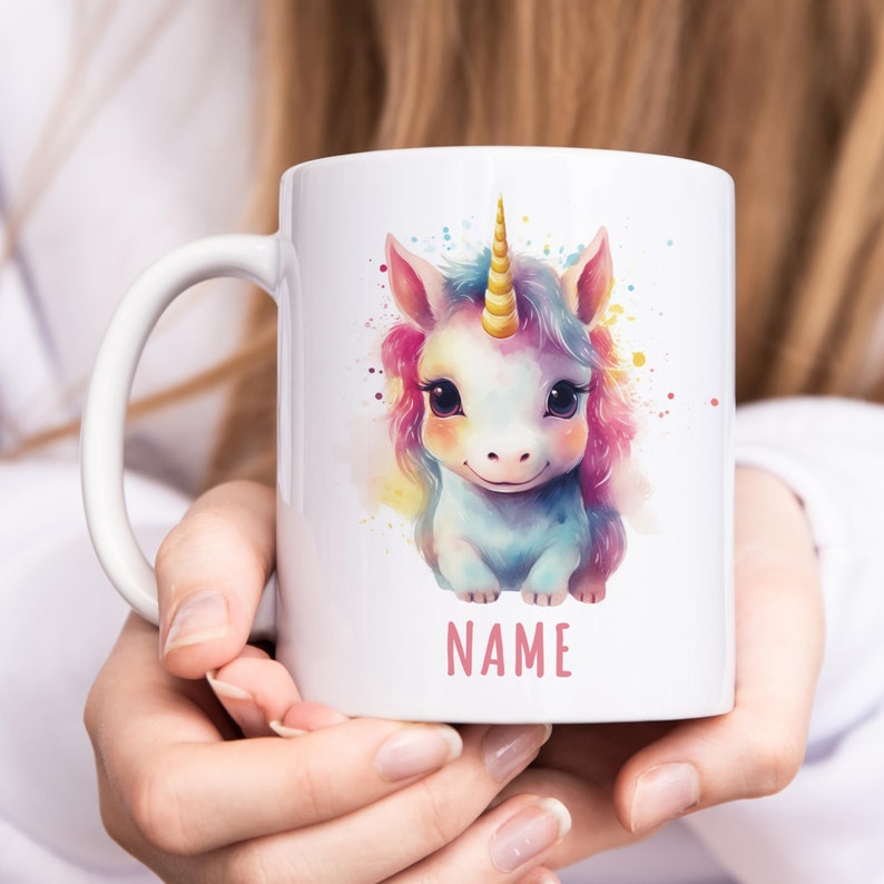 May include: White ceramic mug featuring a colourful illustration of a unicorn with a gold horn. The unicorn is sitting on a watercolour background with pink, blue and yellow splatters. The text "NAME" is printed in pink below the unicorn.